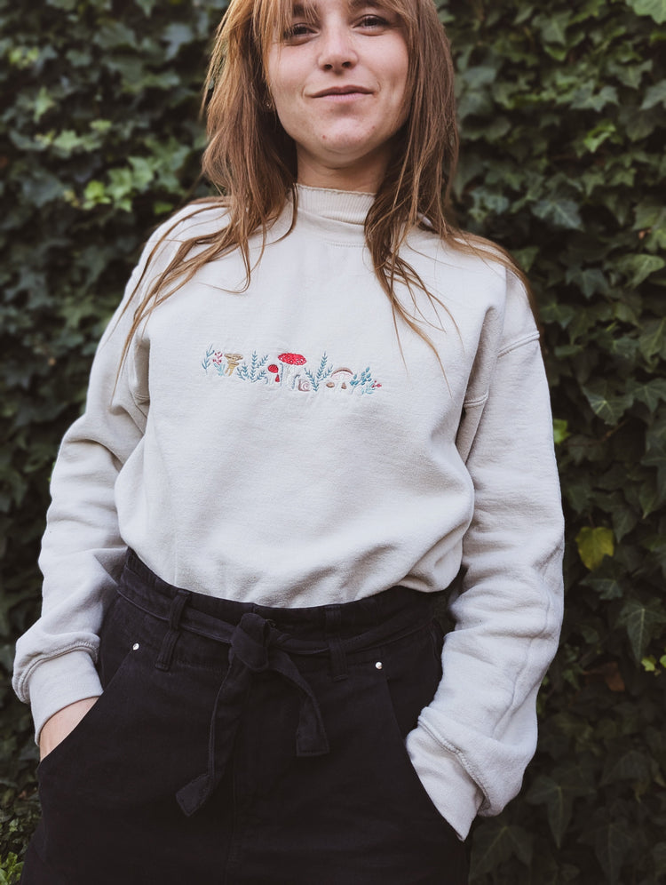 Woman wearing a light-coloured sweatshirt with seasonal embroidered mushrooms and foliage on the chest, standing outdoors against a background of green ivy.