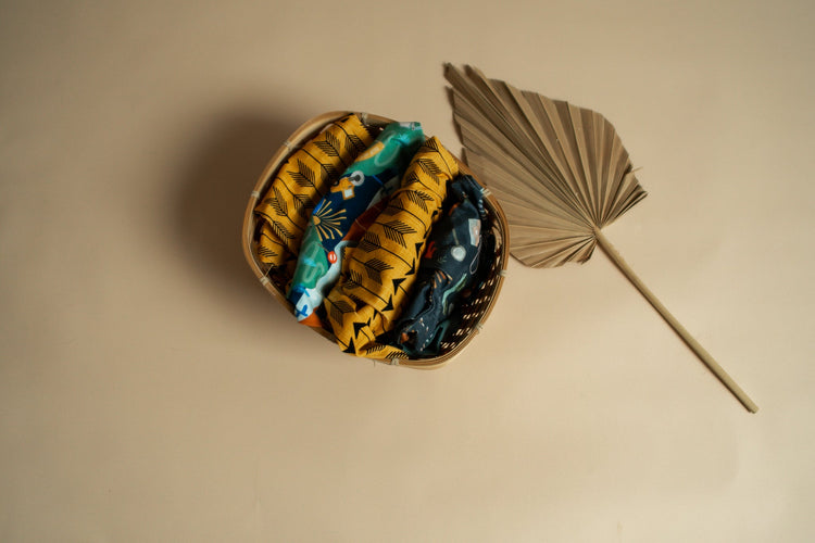 Basket of handmade children’s clothing folded to show vibrant past and unreleased fabric designs, including mustard yellow, teal and construction-themed prints, styled with a dried palm leaf.