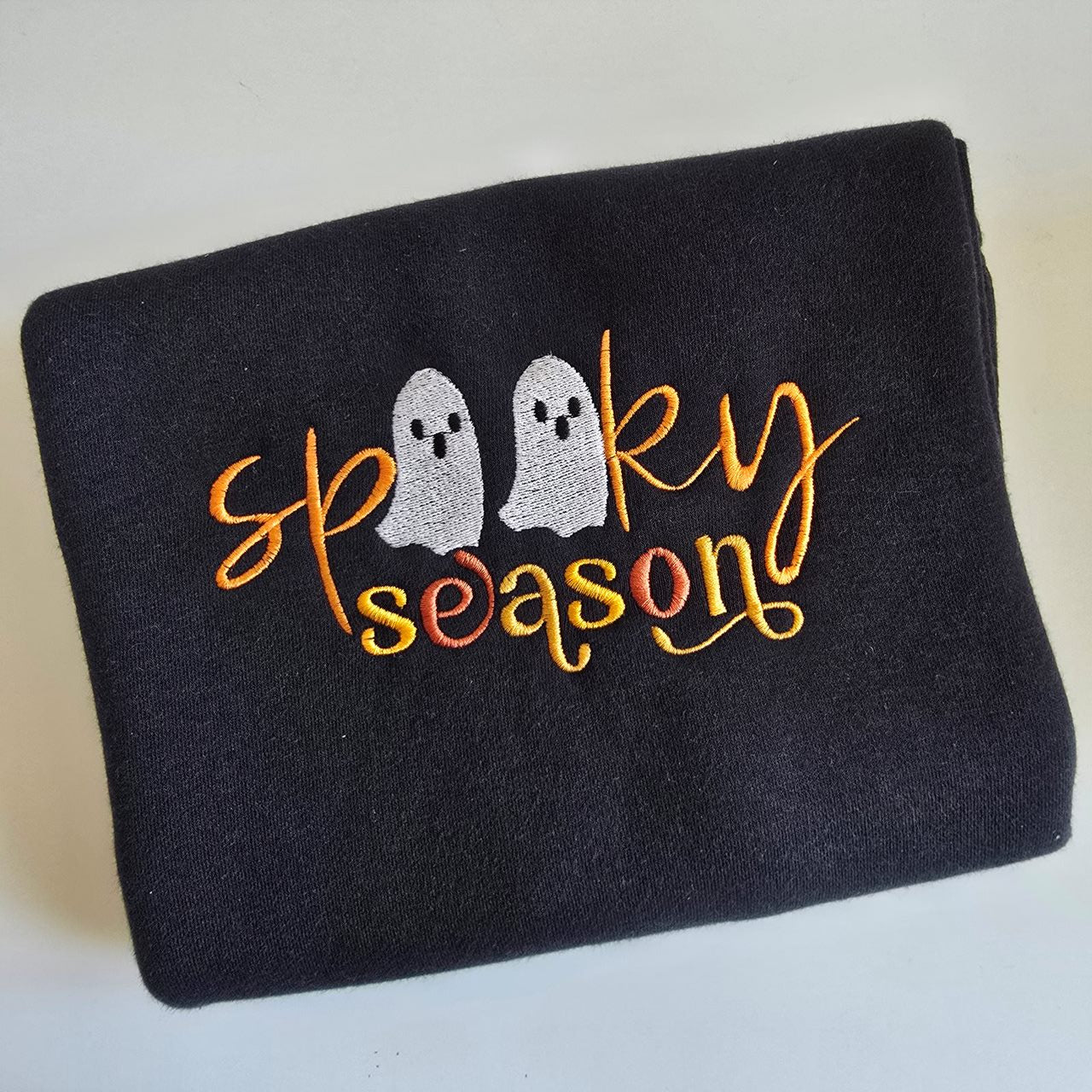 “Spooky season” embroidered in orange and yellow thread with two white ghost characters on black fabric.