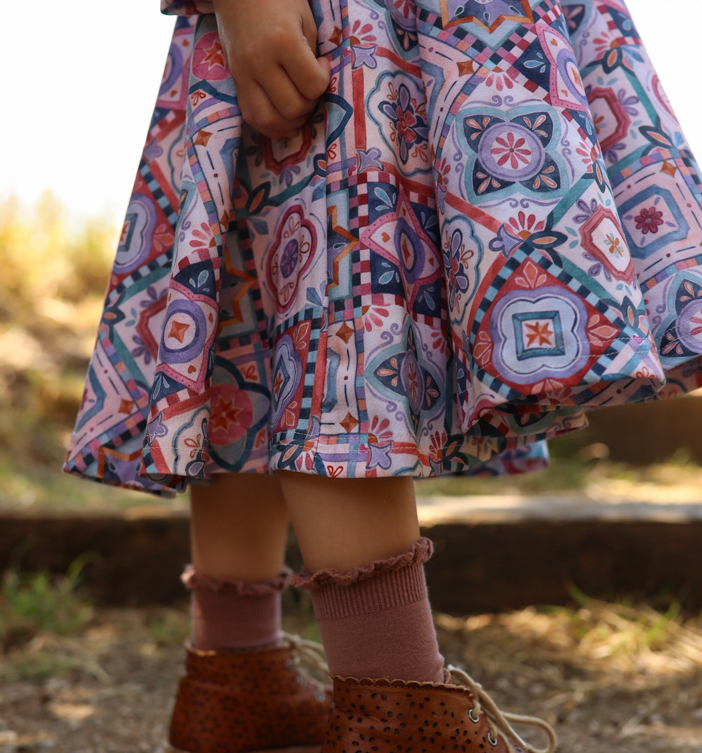 Child wearing a colorful patterned dress and brown boots standing on a wooden log outdoors.