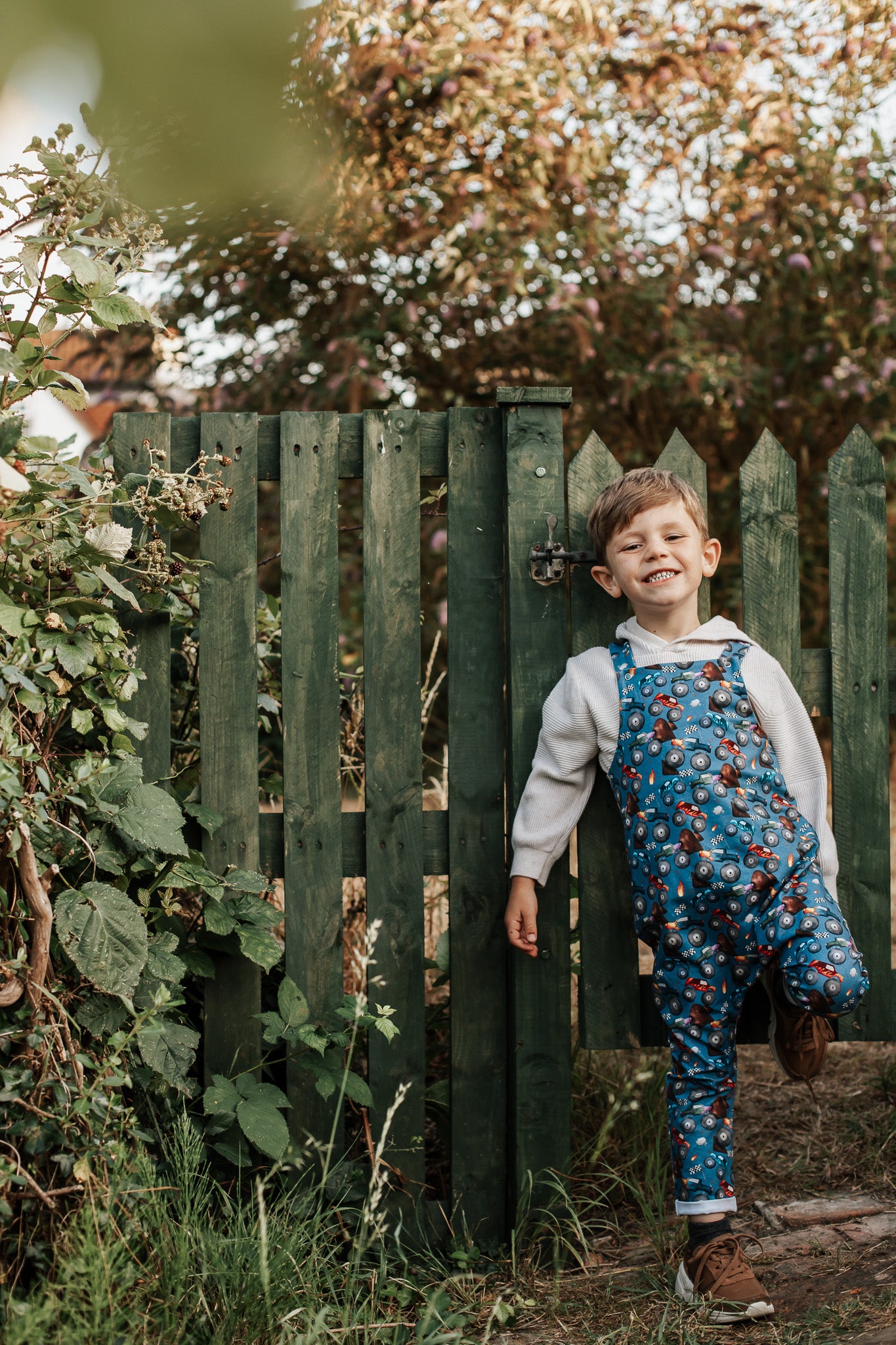 Child in a blue patterned outfit standing in front of a green wooden gate with trees in the background