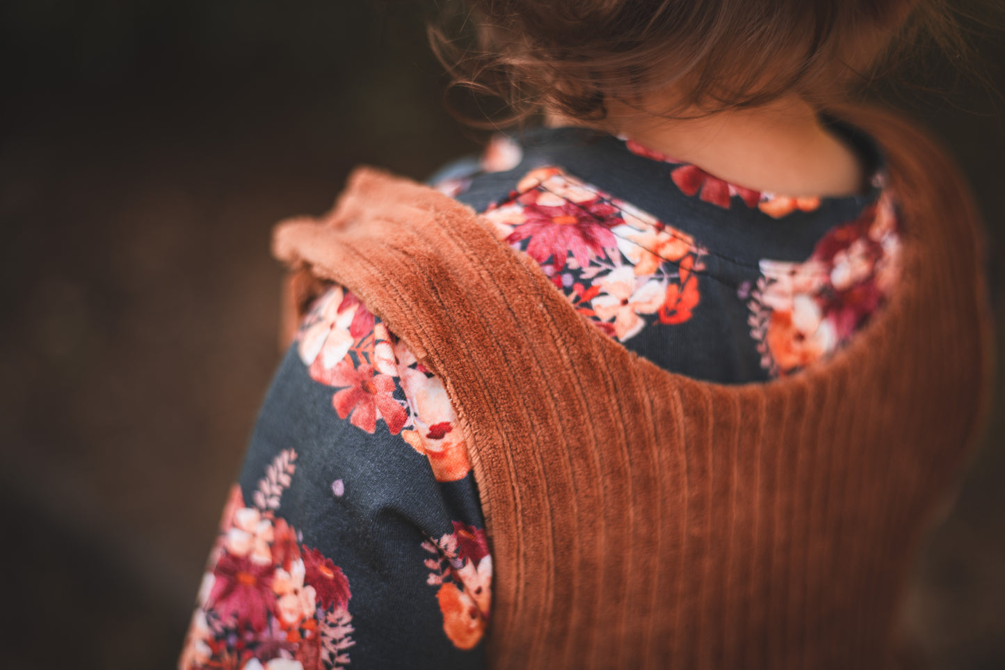 Close-up of a rust chunky corduroy pinafore dress layered over a dark floral top with warm autumn tones.
