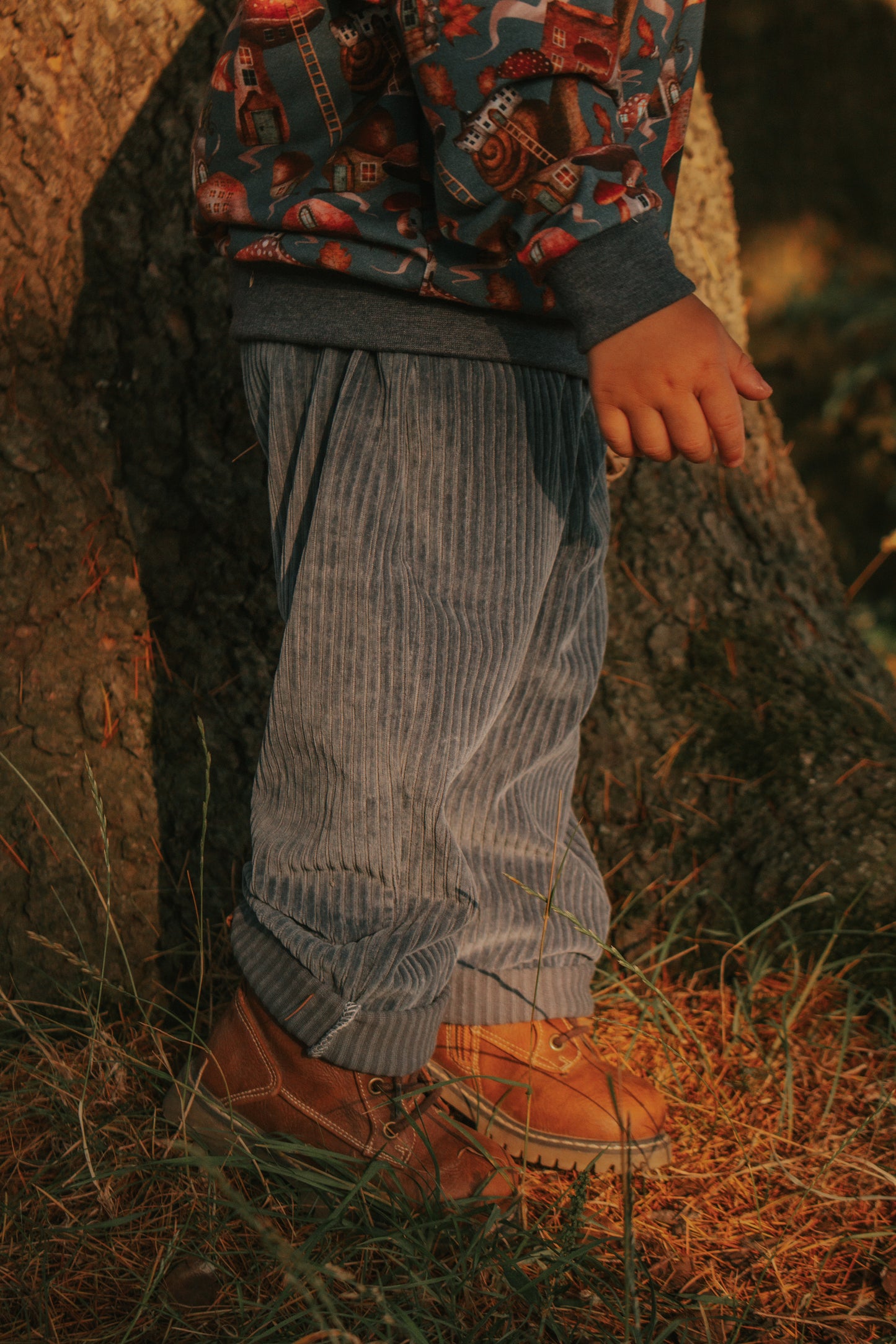Side view of a child wearing jeans blue chunky cord vintage pocket pants with rolled cuffs and brown boots, standing by a tree.