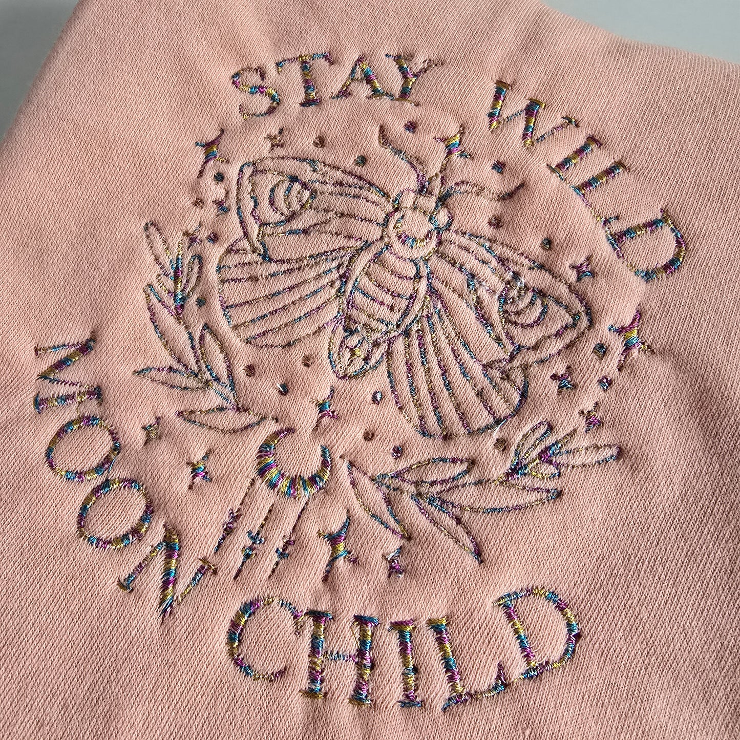 Embroidered “Stay Wild Moon Child” design with a celestial moth and stars in multicoloured thread on pink fabric.