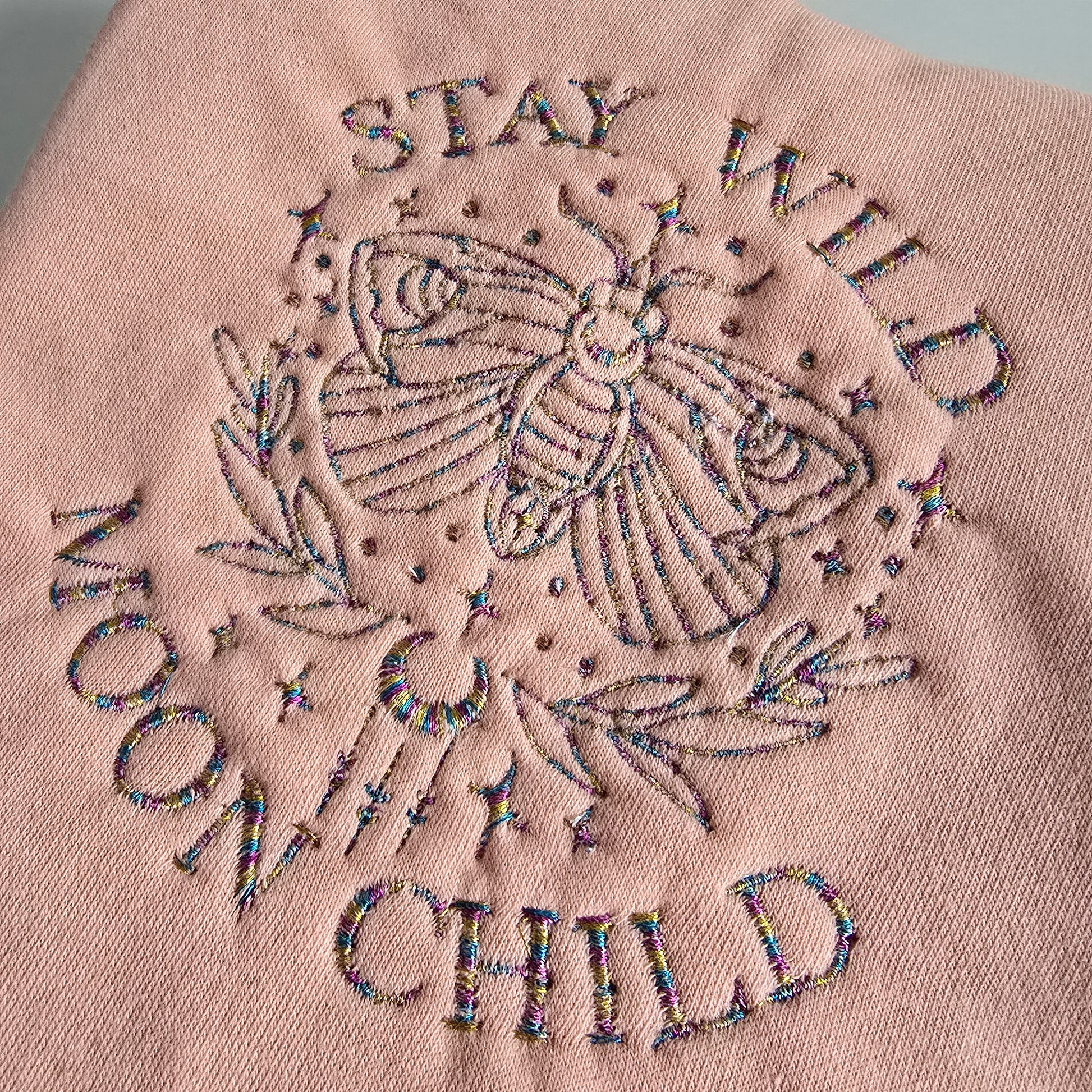 Embroidered “Stay Wild Moon Child” design with a celestial moth and stars in multicoloured thread on pink fabric.