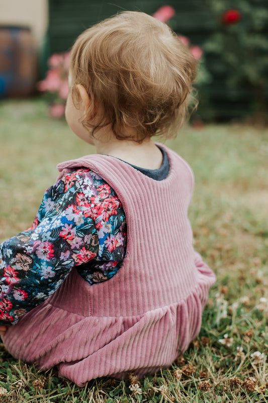Toddler wearing an old lilac chunky corduroy pinafore dress and a bright floral top, sitting on the grass outdoors.