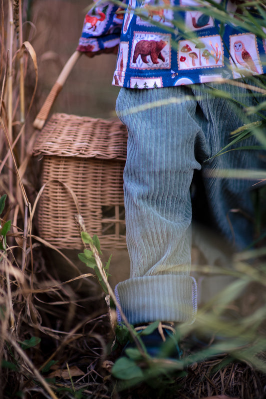 Child in dusty green chunky cord pocket trousers paired with a Wild Stamps top, standing in tall grass holding a wicker basket.