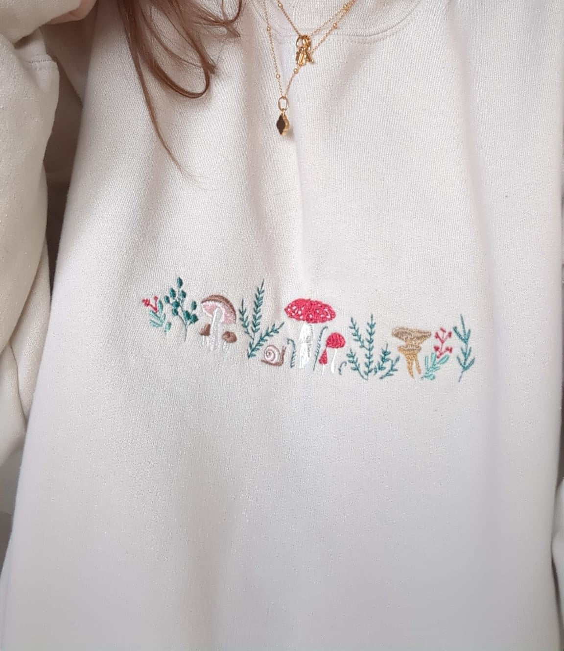 Close-up of a cream sweatshirt with embroidered woodland foliage and mushrooms in soft greens, browns and reds.