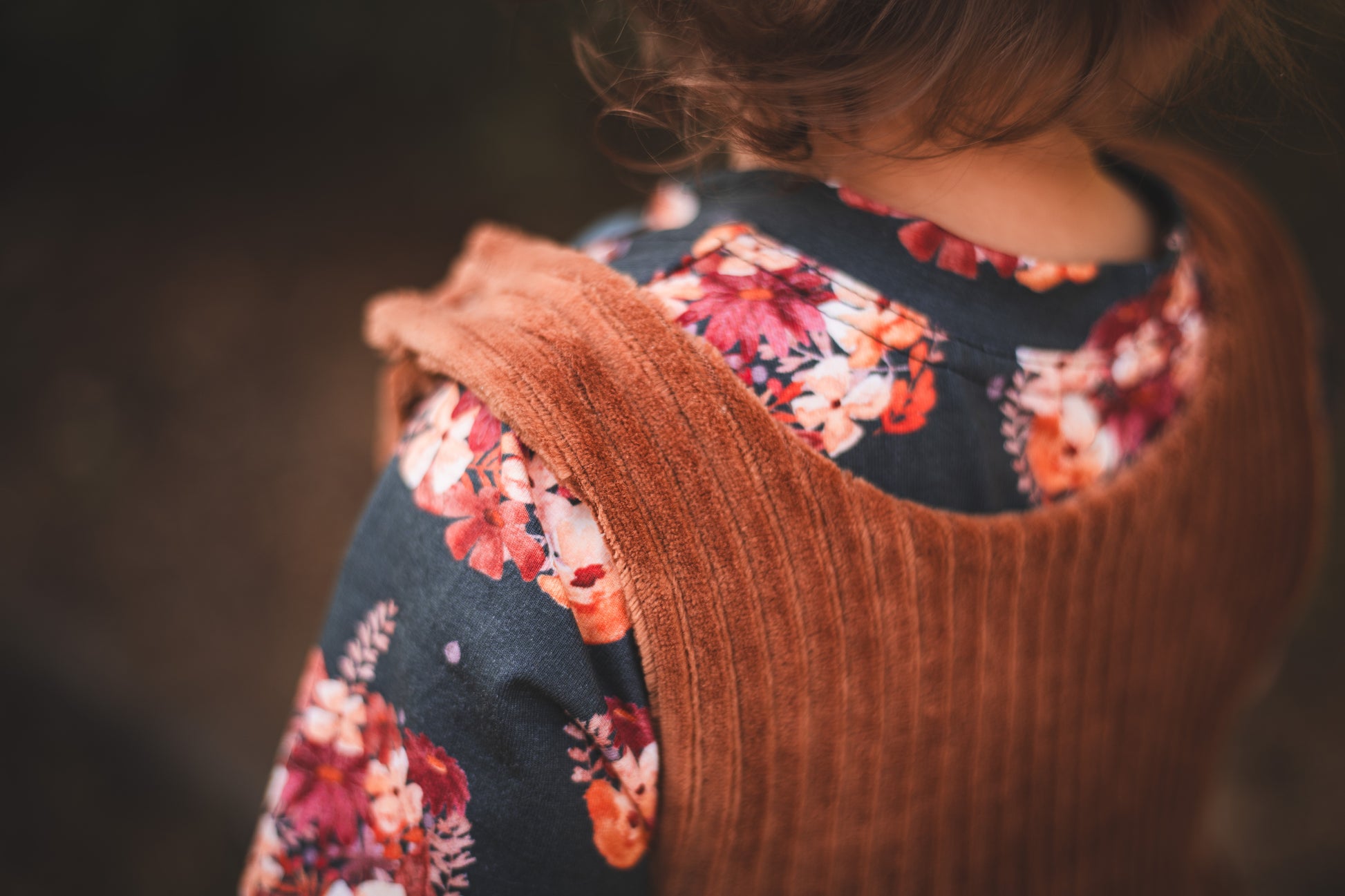Close-up of a rust chunky corduroy pinafore dress layered over a dark floral top with warm autumn tones.
