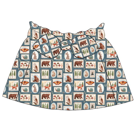Children’s paperbag skirt in Wild Stamps fabric – blue-grey base with animals, mushrooms and bugs in illustrated stamp-style blocks, finished with bow and pockets.