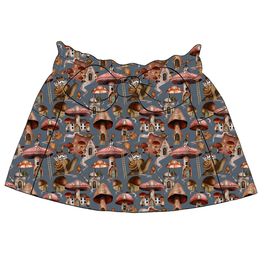 Children’s paperbag skirt in Mushroom Town fabric – blue base with mushroom houses, forest scenes, snails and leafy trees, finished with a bow and pockets.