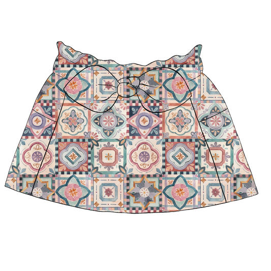 Children’s paperbag skirt in Faro fabric – white base with colourful repeating tiles in pink, mustard, teal and terracotta, with bow and pockets.