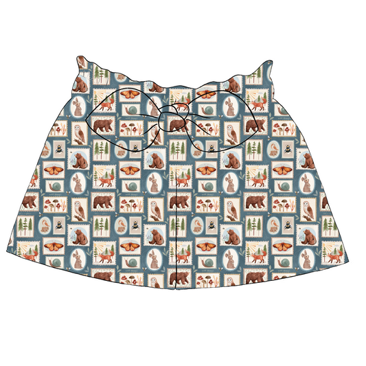 Children’s paperbag shorts in Wild Stamps fabric – blue-grey background with animal portraits, mushrooms and insects in stamp-style blocks, with bow and ruffle waistband.