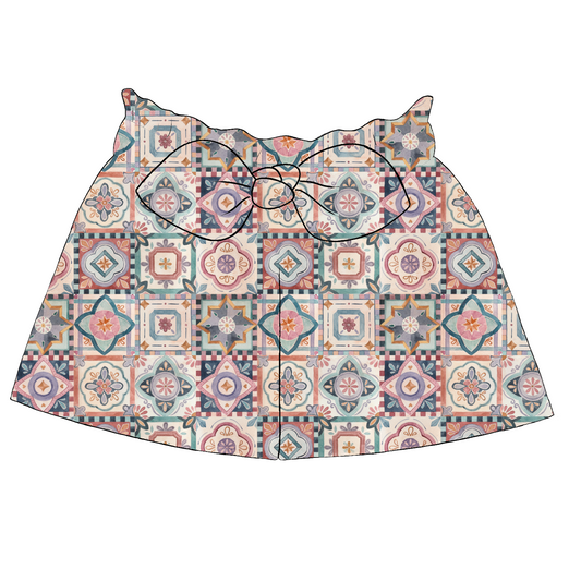 Children’s paperbag shorts in Faro fabric – white base with soft tile pattern in pink, mustard, teal and terracotta, ruffled waistband with bow.