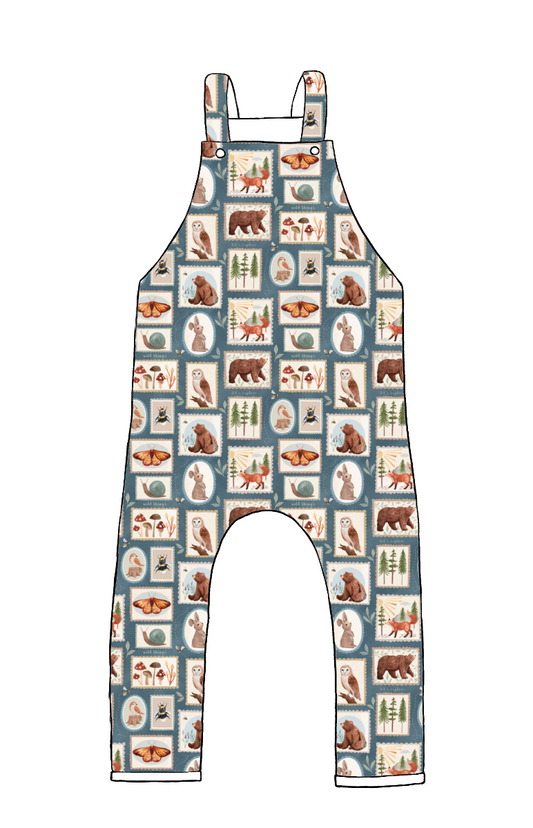 Full-length handmade dungarees in Wild Stamps – blue background with patch-style woodland creatures, insects, and mushrooms in muted colours.