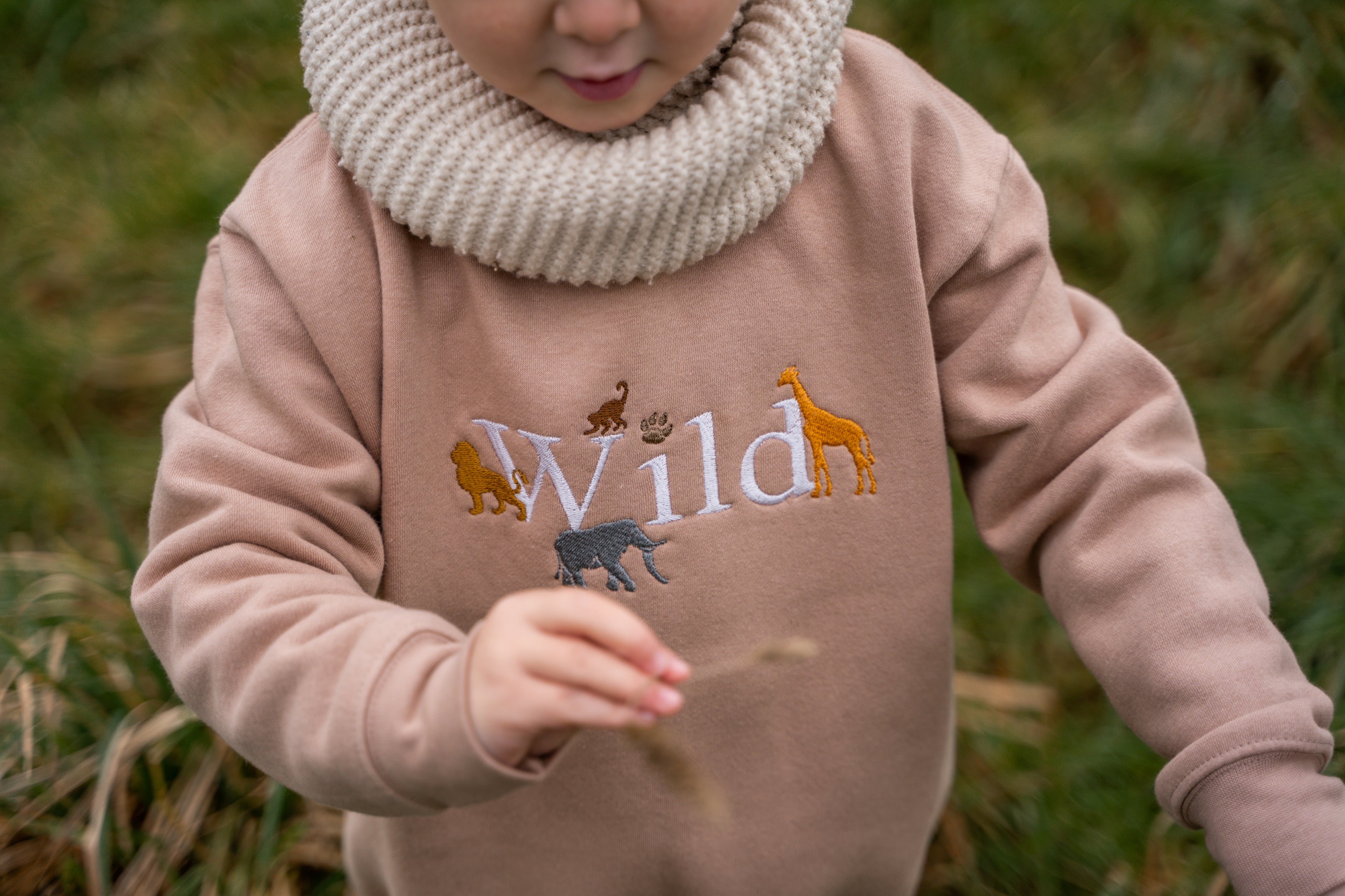 Child wearing a tan sweatshirt with the word “Wild” embroidered alongside safari animals including a lion, giraffe and elephant.